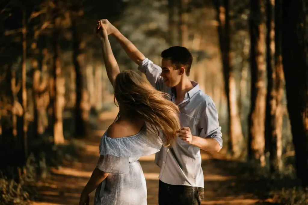 How to show love to another person - A couple dancing together outdoors on a sunlit path through a forest. The scene is warm and romantic, with dappled sunlight filtering through the trees, highlighting the couple's connection and the playful, affectionate nature of their interaction.